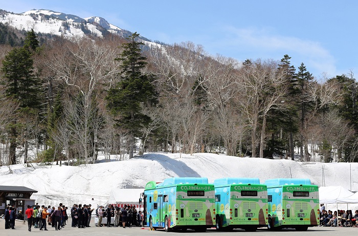 ３台の電気バスと、ダケカンバと針葉樹林帯そして、残雪残る尾瀬のシンボル・標高２３５６ｍの燧ケ岳を奥に望む、記念式典が執り行われた尾瀬御池登山口の様子です。